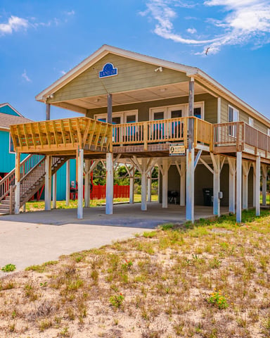 Elevated beach house with yellow deck and wooden pilings, sandy yard, neighboring colorful homes, clear blue sky