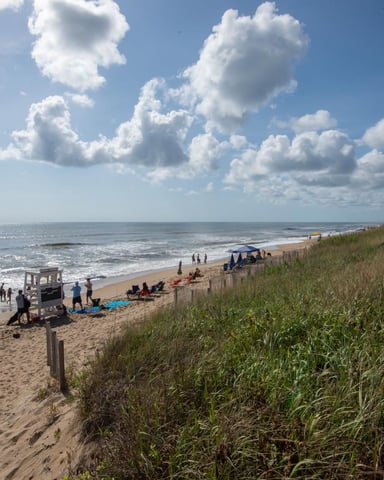 Sandy beach with dune grass, lifeguard tower, beachgoers, and oceanfront houses under blue sky with white clouds