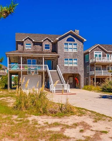 Beachfront shingle-sided house with elevated foundation, white railings, and basketball hoop under clear blue sky.