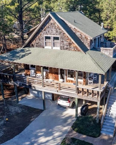 High-angle shot of a cedar-shingled house with a large outdoor deck, parked car underneath, set amidst pine trees and sandy ground, reflecting a rustic, beachside living atmosphere.