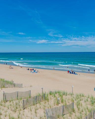 Wide sandy beach with dune fencing and beach grass, scattered beachgoers, turquoise ocean with waves, and clear blue sky
