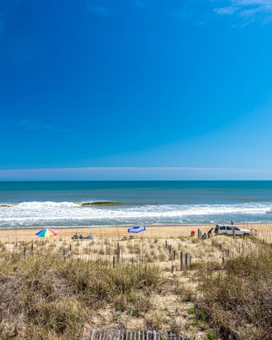 Sandy beach with dune grass, beach umbrellas, and visitors, ocean waves and clear blue sky
