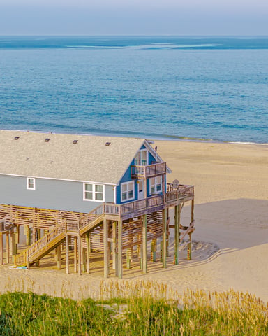 Elevated beach house with blue gable and yellow stilts on sandy shore with ocean view