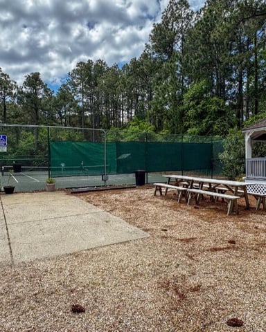 Tennis court with green windscreen, picnic tables, and white gazebo surrounded by tall pine trees