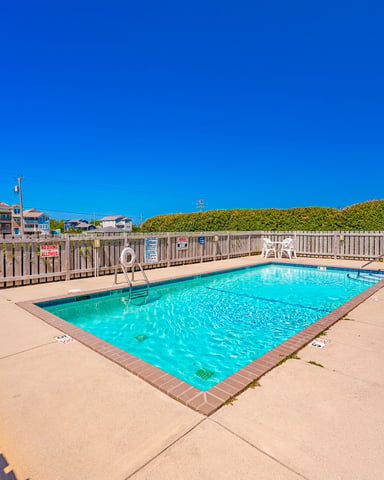 Rectangular swimming pool with turquoise water surrounded by concrete deck, wooden fence, and beach houses under clear blue sky
