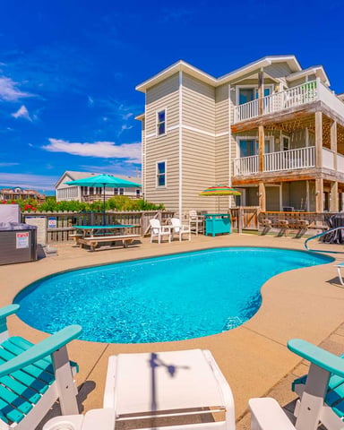 Beachfront vacation rental with swimming pool, lounge chairs, and multi-story house with decks under clear blue sky