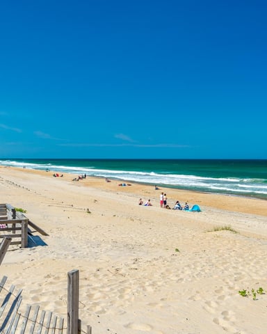 Wide sandy beach with wooden boardwalks, beachgoers, dunes, and oceanfront houses under clear blue sky