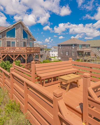 Elevated wooden deck with bench seating overlooking shingle-sided beach houses in a coastal neighborhood under blue sky with white clouds