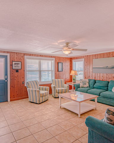 Spacious living room with knotty pine walls, teal upholstered seating, tile flooring, and multiple windows with water views.