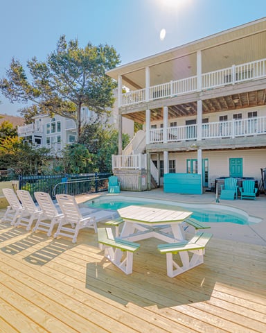 Elevated beach house with turquoise accents, wooden deck, swimming pool, lounge chairs, and surrounding trees under clear sky