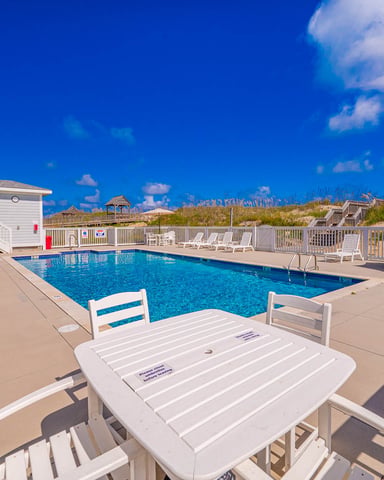 Oceanfront swimming pool with white railings, lounge chairs, and beach houses on sand dunes under blue sky