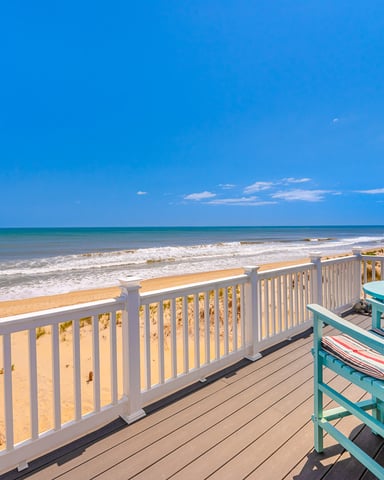 Oceanfront deck with white railing, turquoise Adirondack chairs, sandy beach, and waves under clear blue sky