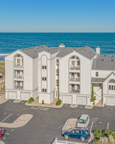 Beachfront condominium building with gray roof and white exterior, ocean and sandy beach visible behind, parking lot in foreground.