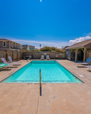 Rectangular swimming pool with turquoise water, concrete deck, lounge chairs, wooden fence, and pool house under clear blue sky