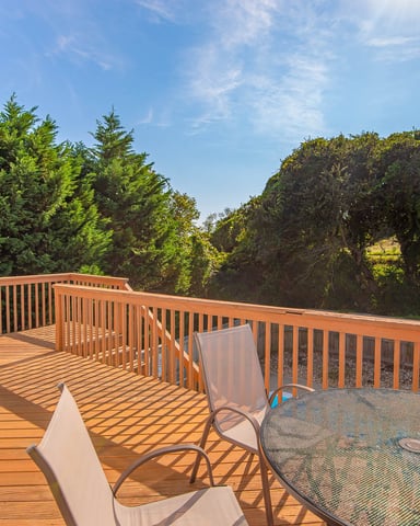 Wooden deck with railing overlooking forested landscape, furnished with lounge chairs and round table