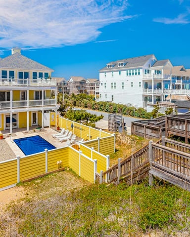 Beachfront vacation home with yellow and white trim, private pool, and wooden deck stairs, surrounded by similar coastal houses under blue sky