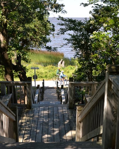 Wooden boardwalk descending through tree-lined path toward water, with person on bicycle visible ahead