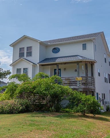Two-story white house with front porch and elevated foundation, surrounded by green lawn and landscaping shrubs.