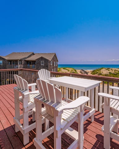 Oceanfront deck with white Adirondack chairs, dining table, and views of beach houses and Atlantic Ocean under clear blue sky