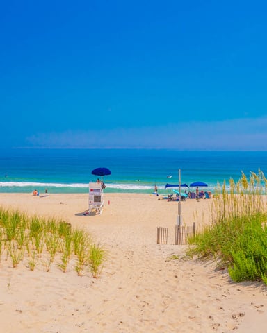 Sandy beach with dune grass, lifeguard stand, beach umbrellas, and swimmers in clear turquoise ocean water under blue sky