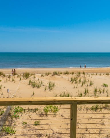 Sandy beach with dune grass and wooden fence, ocean and clear blue sky, swimmers in water