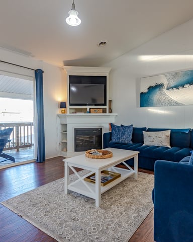 Bright living room with blue sectional sofa, fireplace with TV, white coffee table, and glass doors opening to deck