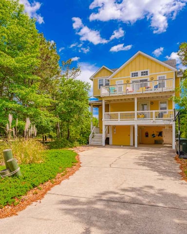 Yellow three-story beach house with white railings and wraparound decks, surrounded by tall trees and a concrete driveway.