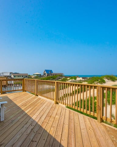 Wooden deck with Adirondack chairs overlooking sand dunes, beach grass, and ocean with coastal homes in distance