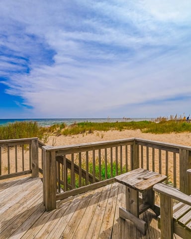 Wooden beach deck with railings overlooking sand dunes, beach grass, and ocean under blue sky