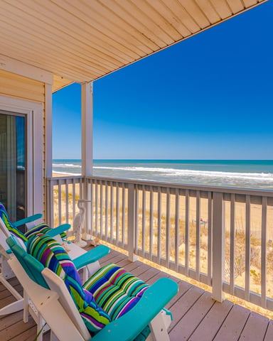 Oceanfront deck with two striped beach chairs overlooking sandy beach, ocean waves, and clear blue sky