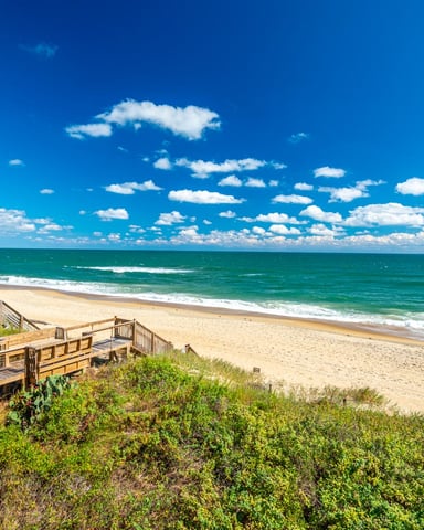 Wooden boardwalk and deck overlooking sandy beach, ocean waves, and blue sky with white clouds