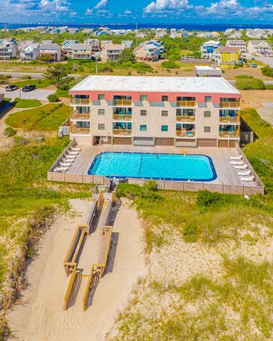 Beachfront condominium complex with swimming pool, wooden boardwalk, and ocean view beyond residential neighborhood.