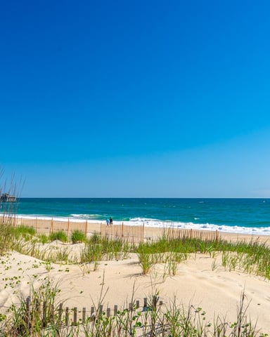 Sandy beach with dune grass and wooden fence, turquoise ocean and white-capped waves under clear blue sky