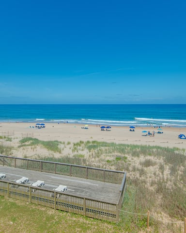 Oceanfront deck with lounge chairs overlooking sandy beach, dunes, and blue Atlantic Ocean with beachgoers and umbrellas