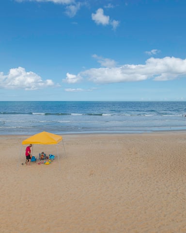 Sandy beach with yellow umbrella tent, swimmers in ocean, and blue sky with white clouds
