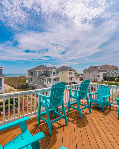Wooden deck with turquoise Adirondack chairs overlooking beach houses and ocean under blue sky