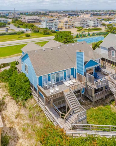 Aerial view of blue beach house with wooden decks and stairs, surrounded by sand dunes and coastal neighborhood with tennis courts and bay beyond.