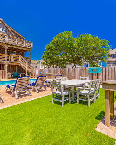 Beachfront vacation home with pool, lounge chairs, dining table, and grill on fenced yard under clear blue sky