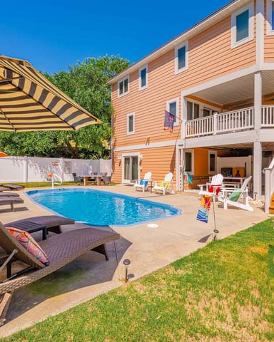 Peach-colored beach house with pool, lounge chairs, striped umbrella, and multi-level decks under clear blue sky