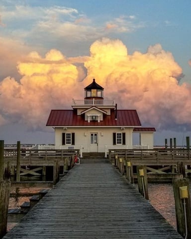White lighthouse with red roof on wooden pier extending into water, dramatic golden clouds at sunset