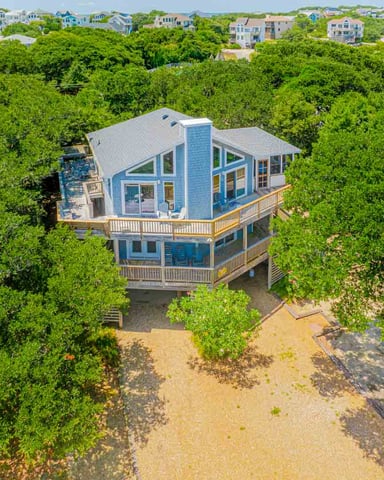 Aerial view of blue beach house on stilts surrounded by dense green vegetation with sandy beach and distant coastal homes