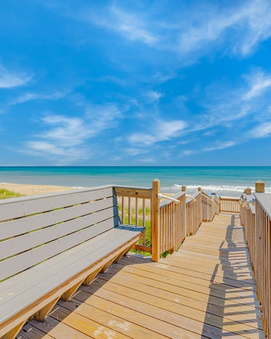 Wooden boardwalk with railings leading to sandy beach, turquoise ocean, and clear blue sky with wispy clouds