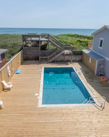 Oceanfront deck with rectangular swimming pool, wooden boardwalk to beach, dune grass, and adjacent rental homes