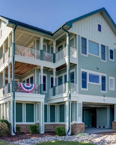 A multi-story pale green house with white railings and shutters, featuring spacious double-decker porches adorned with patriotic bunting, and a well-manicured lawn.