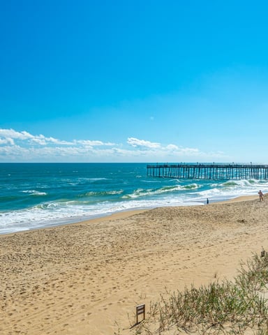 Sandy beach with ocean waves, wooden pier, beachgoers, and gray building under blue sky