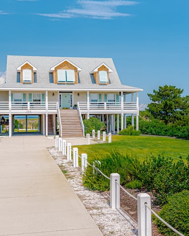 Modern white coastal house with three dormers, elevated on pilings, front porch with railings, concrete driveway, and manicured lawn with white fence.
