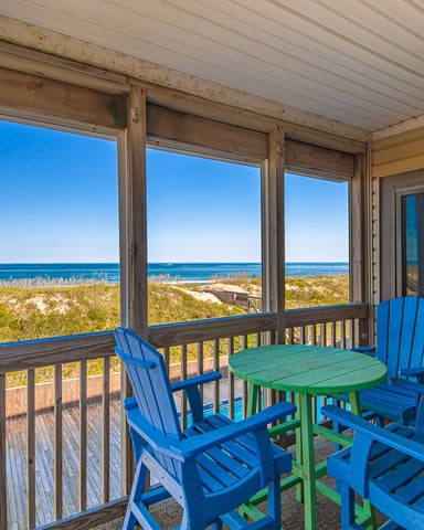Screened porch with blue Adirondack chairs and green table overlooking beach dunes and ocean