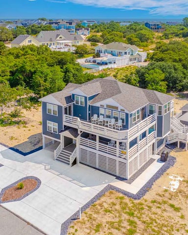 Aerial view of blue and white beachfront house with elevated deck, pool, and sandy lot surrounded by trees and neighboring homes.
