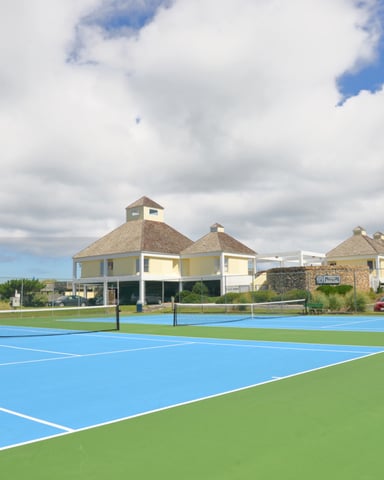 Blue and green tennis courts with elevated beach houses and parked cars in background under cloudy sky