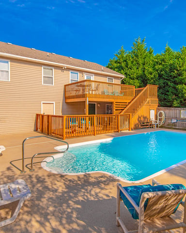 Tan two-story house with wooden deck and rectangular swimming pool, lounge chairs, and pink flowering shrubs in fenced backyard